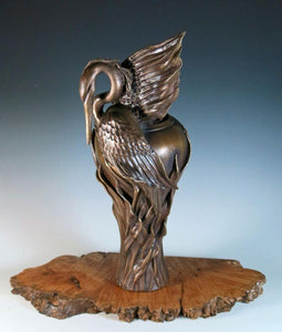 Bronze sculpture of a hunting heron with wings back poised on a reed covered vase. Set on a base of redwood burl with a natural edge.