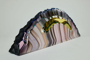 Kiln formed glass sculpture in soft pink, lavender, purple, emerald and gold ribbons of color in an arched shaped piece. The top arched edge is textured with smooth, flat sides and bottom.